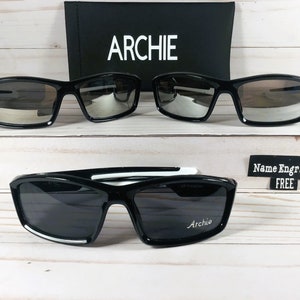 May include: Two pairs of black sunglasses with mirrored lenses. The top pair has a black frame. The bottom pair has a white stripe on the frame. The bottom pair has the word "Archie" engraved on the right lens. A sign reads "Name Engraved FREE".