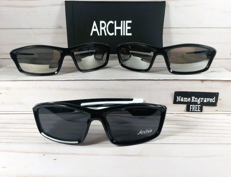 May include: Two pairs of black sunglasses with mirrored lenses. The top pair has a black frame. The bottom pair has a white stripe on the frame. The bottom pair has the word "Archie" engraved on the right lens. A sign reads "Name Engraved FREE".