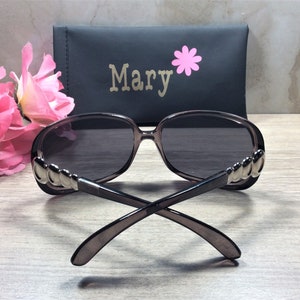 Puede incluir: Un par de gafas de sol de color gris oscuro con detalles plateados en las patillas. Las gafas de sol est&aacute;n dobladas y sobre una superficie de madera. Las gafas de sol est&aacute;n en una funda negra con el nombre "Mary" impreso en ella.
