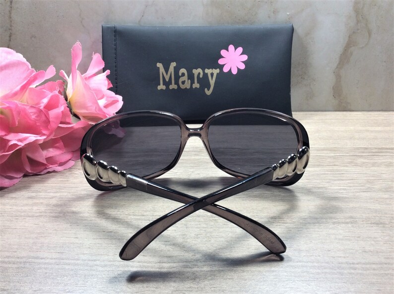 Puede incluir: Un par de gafas de sol de color gris oscuro con detalles plateados en las patillas. Las gafas de sol est&aacute;n dobladas y sobre una superficie de madera. Las gafas de sol est&aacute;n en una funda negra con el nombre "Mary" impreso en ella.