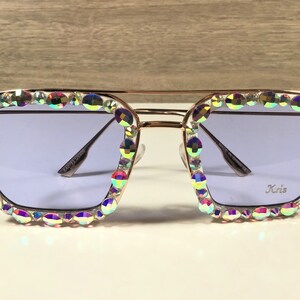 Peut inclure: Lunettes de soleil dor&eacute;es avec une monture carr&eacute;e et des strass iris&eacute;s autour des verres. Les verres sont de couleur violet clair. Les lunettes de soleil portent l'inscription "Kris" sur le verre droit.