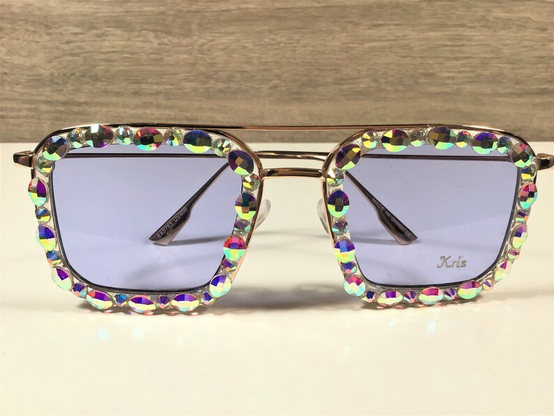Peut inclure: Lunettes de soleil dor&eacute;es avec une monture carr&eacute;e et des strass iris&eacute;s autour des verres. Les verres sont de couleur violet clair. Les lunettes de soleil portent l'inscription "Kris" sur le verre droit.