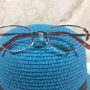 Pu&ograve; includere: Occhiali con montatura argentata, aste rosse e decorazioni rosa. Le lenti sono ovali e le aste riportano la scritta "UV Protection". Gli occhiali sono appoggiati su un cappello blu.