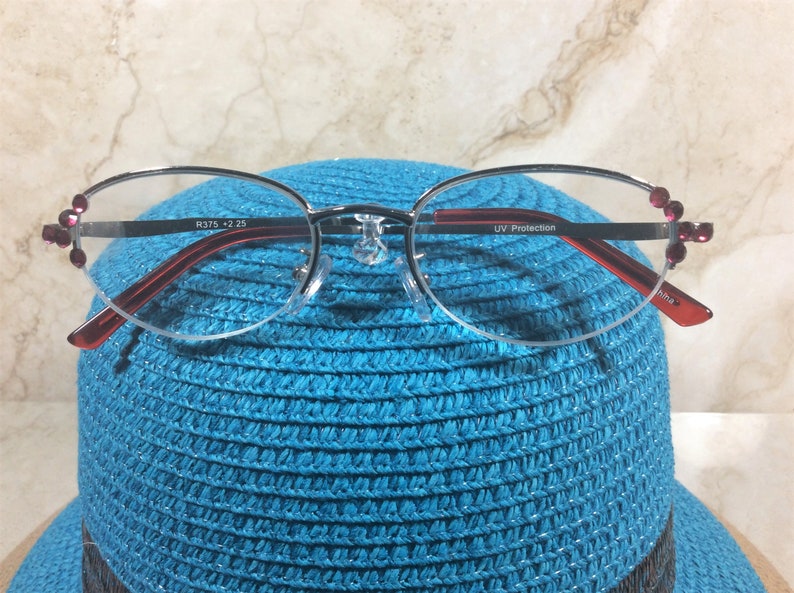 Pu&ograve; includere: Occhiali con montatura argentata, aste rosse e decorazioni rosa. Le lenti sono ovali e le aste riportano la scritta "UV Protection". Gli occhiali sono appoggiati su un cappello blu.
