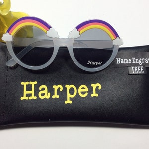 Puede incluir: Gafas de sol redondas de pl&aacute;stico blanco con un dise&ntilde;o de arco&iacute;ris y nubes. Las gafas est&aacute;n en una funda negra con el nombre "Harper" impreso en letras amarillas.