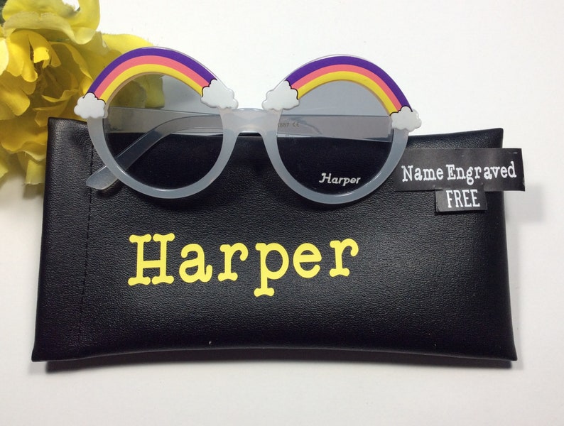 Puede incluir: Gafas de sol redondas de pl&aacute;stico blanco con un dise&ntilde;o de arco&iacute;ris y nubes. Las gafas est&aacute;n en una funda negra con el nombre "Harper" impreso en letras amarillas.