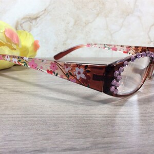 May include: Brown-framed spectacles with floral patterned arms and clear lenses. The frames are decorated with pink rhinestones at the corners. The arms feature a design of pink and white flowers on a cream background.