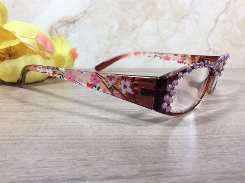 May include: Brown-framed spectacles with floral patterned arms and clear lenses. The frames are decorated with pink rhinestones at the corners. The arms feature a design of pink and white flowers on a cream background.