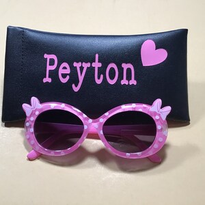 May include: Pink polka dot sunglasses with a bow detail on each side, in a black case with the name Peyton and a pink heart.