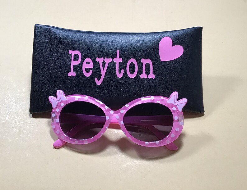 May include: Pink polka dot sunglasses with a bow detail on each side, in a black case with the name Peyton and a pink heart.