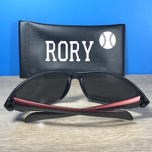 Puede incluir: Gafas de sol negras con detalles rojos y una funda negra con el nombre "RORY" y un gr&aacute;fico de b&eacute;isbol blanco.