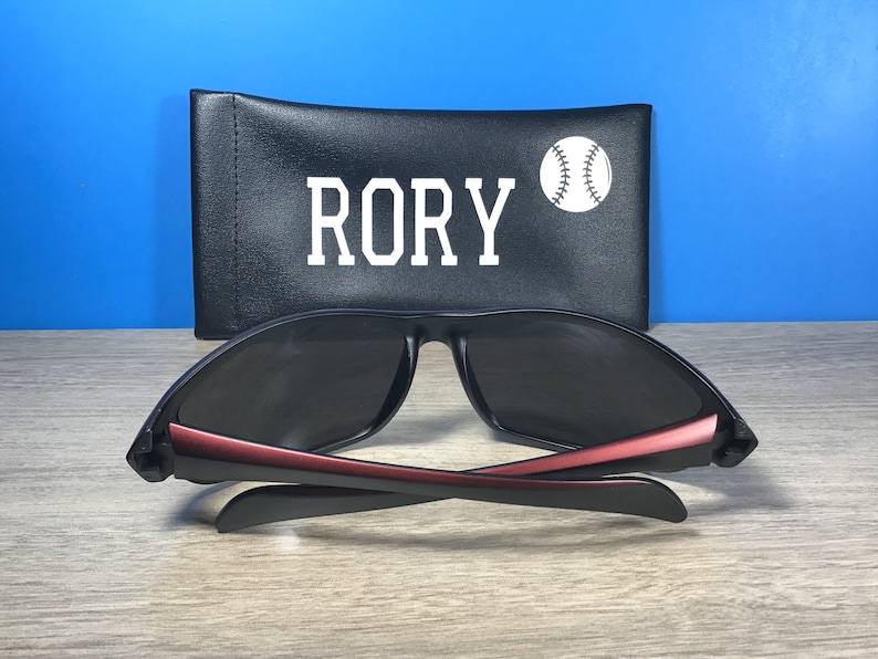 Puede incluir: Gafas de sol negras con detalles rojos y una funda negra con el nombre "RORY" y un gr&aacute;fico de b&eacute;isbol blanco.