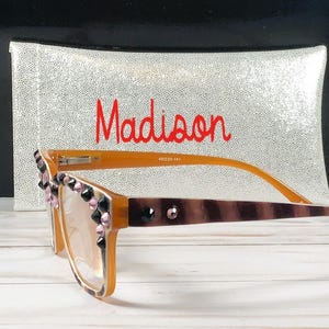 Op de afbeelding: Bruine en bruine bril met decoratieve zwarte en roze versieringen. De bril heeft een gestreept patroon op de pootjes en wordt geleverd met een zilveren etui met de naam "Madison" in het rood.