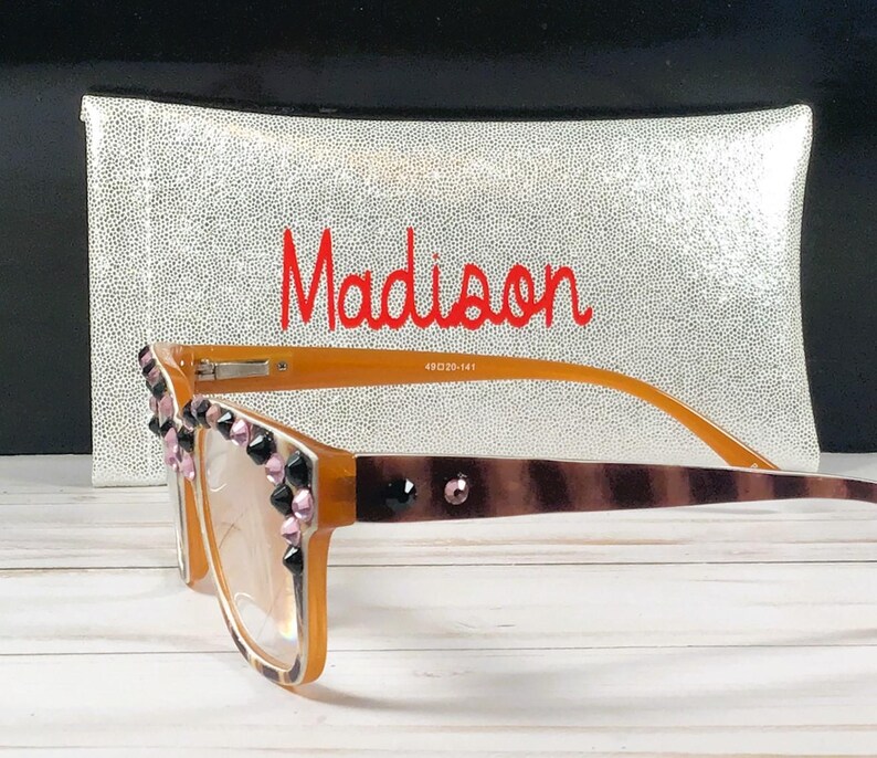 Op de afbeelding: Bruine en bruine bril met decoratieve zwarte en roze versieringen. De bril heeft een gestreept patroon op de pootjes en wordt geleverd met een zilveren etui met de naam "Madison" in het rood.