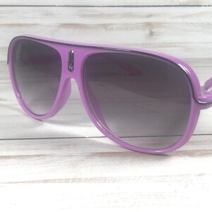 Puede incluir: Gafas de sol de color lila con lentes degradadas oscuras. Las gafas tienen una montura gruesa y una forma de aviador cl&aacute;sica. Las patillas est&aacute;n unidas a la montura con una peque&ntilde;a bisagra. Las lentes son oscuras, proporcionando sombra y estilo.