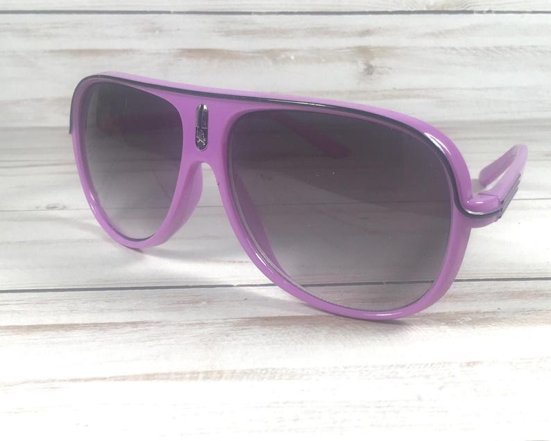 Puede incluir: Gafas de sol de color lila con lentes degradadas oscuras. Las gafas tienen una montura gruesa y una forma de aviador cl&aacute;sica. Las patillas est&aacute;n unidas a la montura con una peque&ntilde;a bisagra. Las lentes son oscuras, proporcionando sombra y estilo.