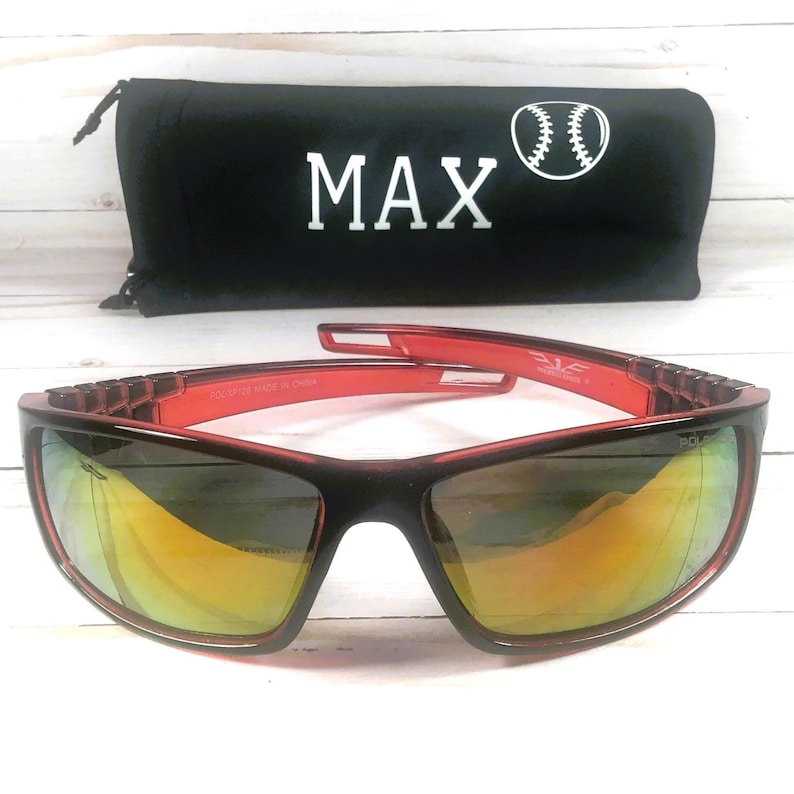May include: Black and red sunglasses with mirrored lenses. The sunglasses are paired with a black carrying case with the word "MAX" in white and a baseball graphic. The sunglasses have a sporty design.