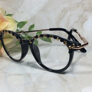 May include: Black cat-eye spectacles with clear lenses, featuring gold and black decorative elements along the top rims. The frames have a black front and gold-toned temples. The glasses are displayed against a soft background with floral accents.