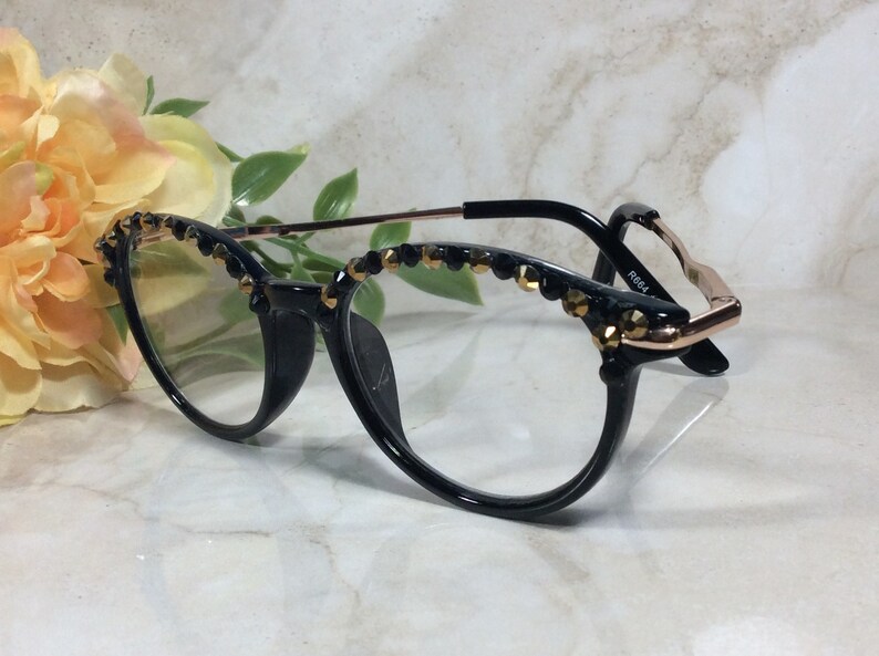May include: Black cat-eye spectacles with clear lenses, featuring gold and black decorative elements along the top rims. The frames have a black front and gold-toned temples. The glasses are displayed against a soft background with floral accents.
