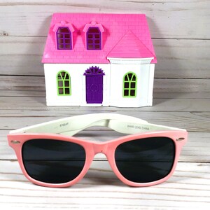 Op de afbeelding: Roze plastic zonnebril met zwarte glazen en witte pootjes. De zonnebril ligt op een houten oppervlak voor een roze en wit speelgoedhuis.