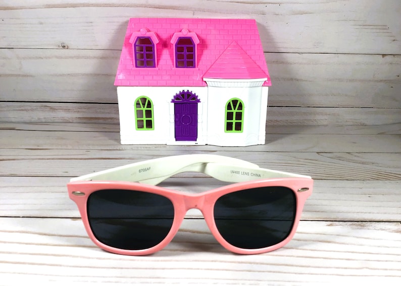 Op de afbeelding: Roze plastic zonnebril met zwarte glazen en witte pootjes. De zonnebril ligt op een houten oppervlak voor een roze en wit speelgoedhuis.