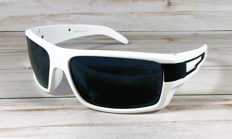 Pu&ograve; includere: Occhiali da sole bianchi con lenti scure. Gli occhiali hanno una montatura spessa e curva e un design elegante. Le lenti sono scure, offrendo protezione solare. Sono appoggiati su una superficie di legno chiaro.