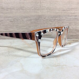 Puede incluir: Gafas con un estampado de rayas de cebra marr&oacute;n y blanco en las patillas y los bordes de la montura. Las monturas son de color marr&oacute;n claro y est&aacute;n adornadas con pedrer&iacute;a transparente. Las lentes son transparentes.