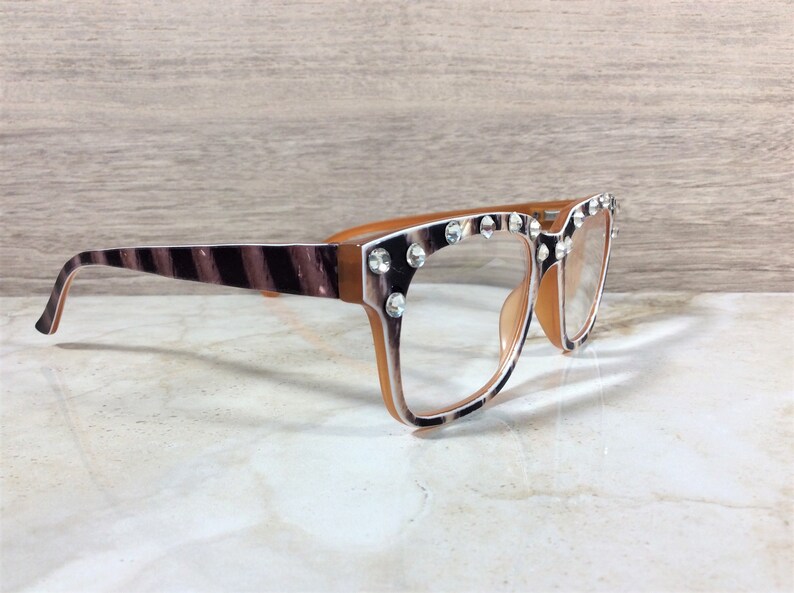 Puede incluir: Gafas con un estampado de rayas de cebra marr&oacute;n y blanco en las patillas y los bordes de la montura. Las monturas son de color marr&oacute;n claro y est&aacute;n adornadas con pedrer&iacute;a transparente. Las lentes son transparentes.