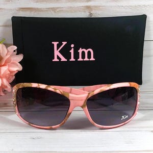 May include: Pink camo sunglasses with rhinestones and a black fabric case with the name "Kim" embroidered in pink.