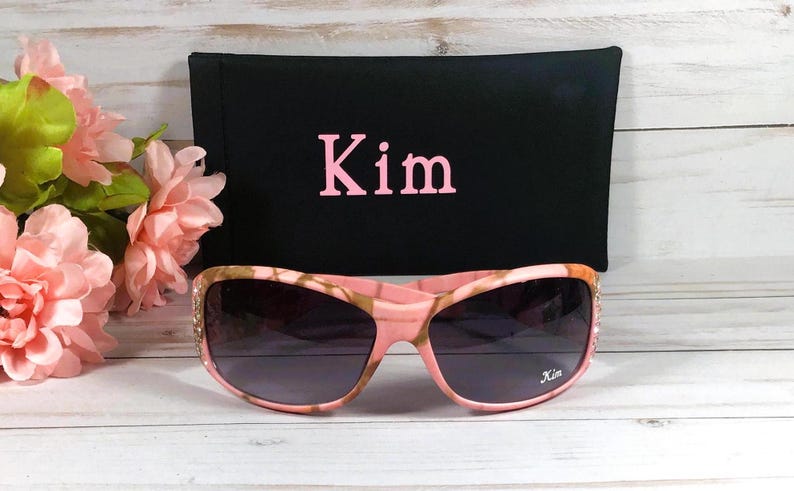 May include: Pink camo sunglasses with rhinestones and a black fabric case with the name "Kim" embroidered in pink.
