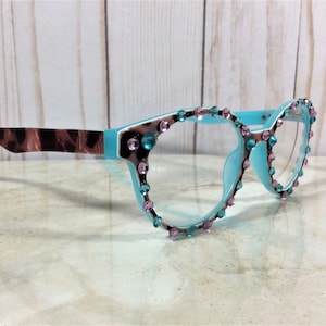 May include: A pair of eyeglasses with a light blue frame, adorned with pink and teal gemstones. The temples feature a brown and tan animal print. The glasses are set against a light wood background.