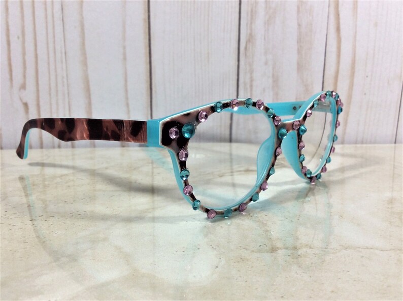 May include: A pair of eyeglasses with a light blue frame, adorned with pink and teal gemstones. The temples feature a brown and tan animal print. The glasses are set against a light wood background.