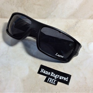 May include: Black sunglasses with a sporty design. The sunglasses have dark lenses and a matte black frame. The name "Layne" is engraved on the lens. A small sign reads "Name Engraved FREE".