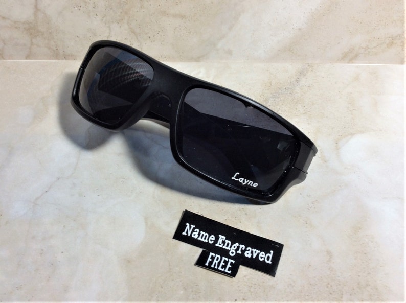 May include: Black sunglasses with a sporty design. The sunglasses have dark lenses and a matte black frame. The name "Layne" is engraved on the lens. A small sign reads "Name Engraved FREE".