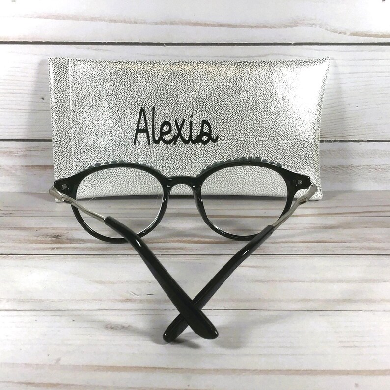 Puede incluir: Gafas de montura negra con lentes transparentes colocadas frente a un estuche rectangular con purpurina plateada. El estuche presenta el nombre Alexia impreso en cursiva negra en el centro. Las gafas tienen patillas met&aacute;licas finas.