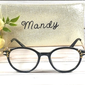 May include: Black-framed eyeglasses with gold accents and a gold-colored case with the name "Mandy" in script. The glasses are in focus, with a blurred background of a floral arrangement and a textured case.