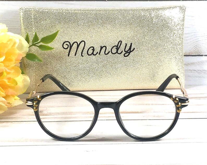 May include: Black-framed eyeglasses with gold accents and a gold-colored case with the name "Mandy" in script. The glasses are in focus, with a blurred background of a floral arrangement and a textured case.