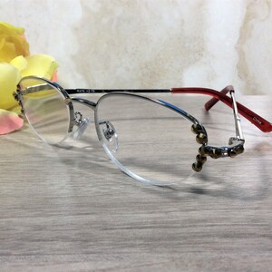 Puede incluir: Gafas con montura plateada, lentes transparentes y patillas rojas. Las monturas tienen adornos decorativos en las esquinas inferiores. El texto "RS75 +2.75" est&aacute; impreso en la montura. Las gafas est&aacute;n sobre una superficie de madera clara.
