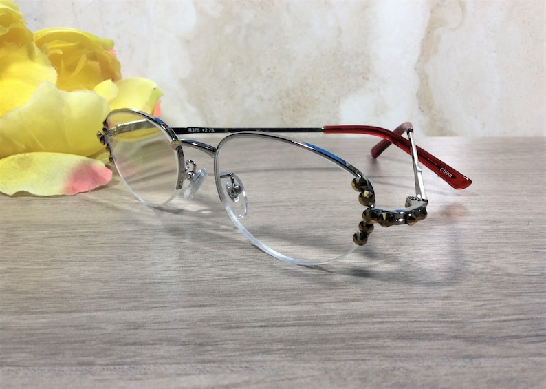 Puede incluir: Gafas con montura plateada, lentes transparentes y patillas rojas. Las monturas tienen adornos decorativos en las esquinas inferiores. El texto "RS75 +2.75" est&aacute; impreso en la montura. Las gafas est&aacute;n sobre una superficie de madera clara.