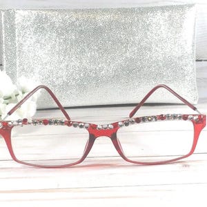 May include: Red-framed eyeglasses with decorative rhinestones along the top rim. The glasses have a rectangular shape and are set against a silver, textured background. The overall style is fashionable and eye-catching.