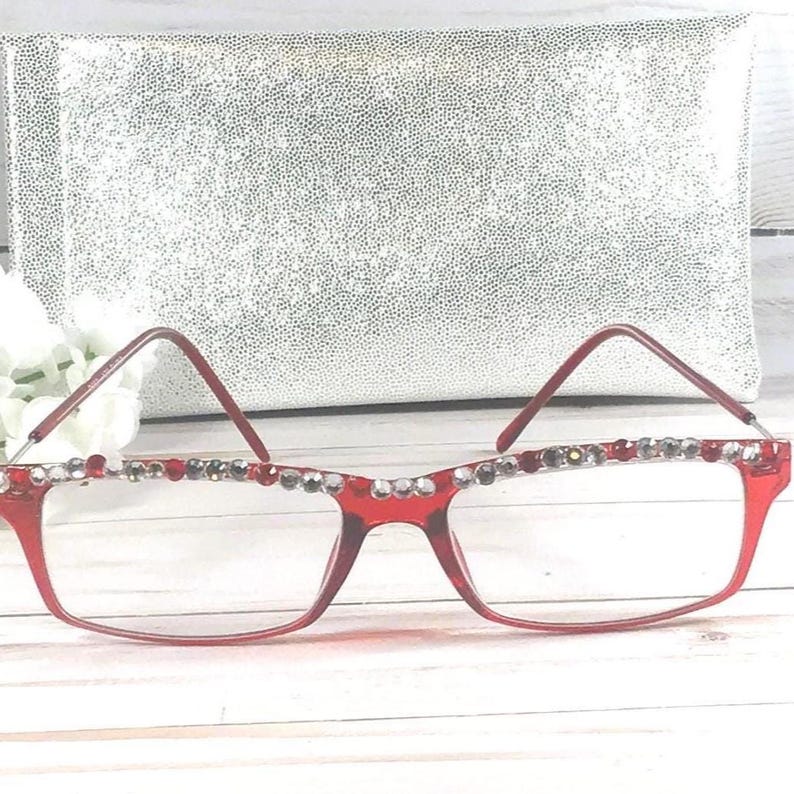 May include: Red-framed eyeglasses with decorative rhinestones along the top rim. The glasses have a rectangular shape and are set against a silver, textured background. The overall style is fashionable and eye-catching.