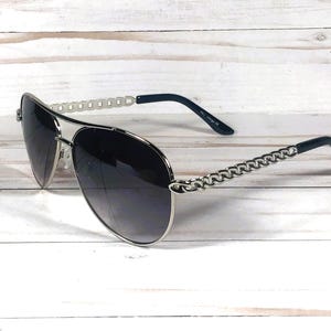 Peut inclure: Lunettes de soleil aviateur argent&eacute;es avec des verres gris fonc&eacute; et un d&eacute;tail de cha&icirc;ne sur les branches.