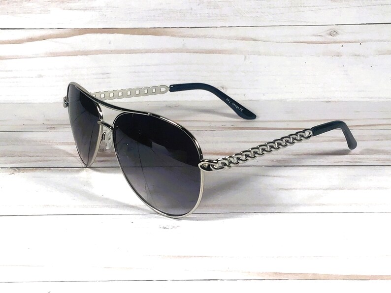Peut inclure: Lunettes de soleil aviateur argent&eacute;es avec des verres gris fonc&eacute; et un d&eacute;tail de cha&icirc;ne sur les branches.