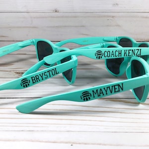 Puede incluir: Tres pares de gafas de sol de color turquesa claro con lentes oscuras. Cada patilla presenta un icono de voleibol negro seguido de un nombre o t&iacute;tulo: COACH KENZI, BRYSTOL y MAYVEN. Las monturas son de material pl&aacute;stico mate.