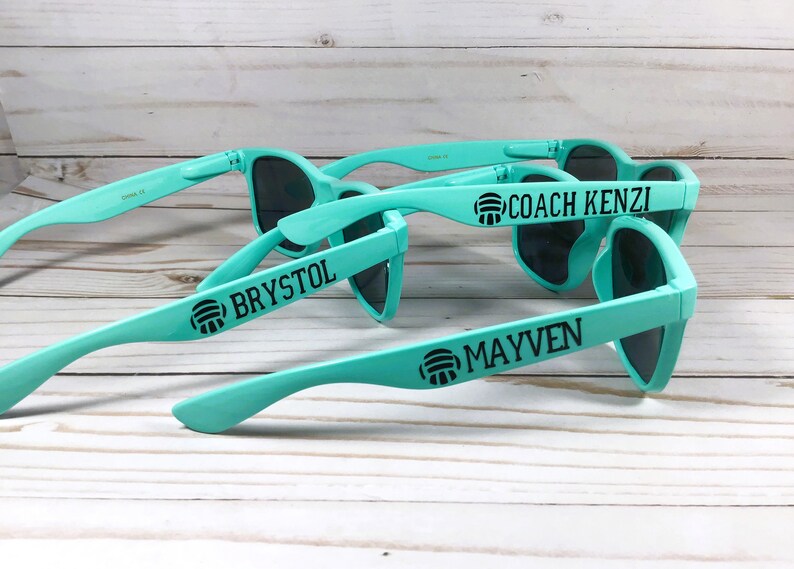 Puede incluir: Tres pares de gafas de sol de color turquesa claro con lentes oscuras. Cada patilla presenta un icono de voleibol negro seguido de un nombre o t&iacute;tulo: COACH KENZI, BRYSTOL y MAYVEN. Las monturas son de material pl&aacute;stico mate.