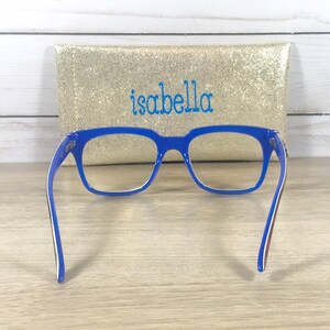 Puede incluir: Gafas azules con montura rectangular frente a un estuche dorado con lentejuelas bordado con el nombre "isabella" en azul. Las gafas tienen detalles blancos y marrones en las patillas.