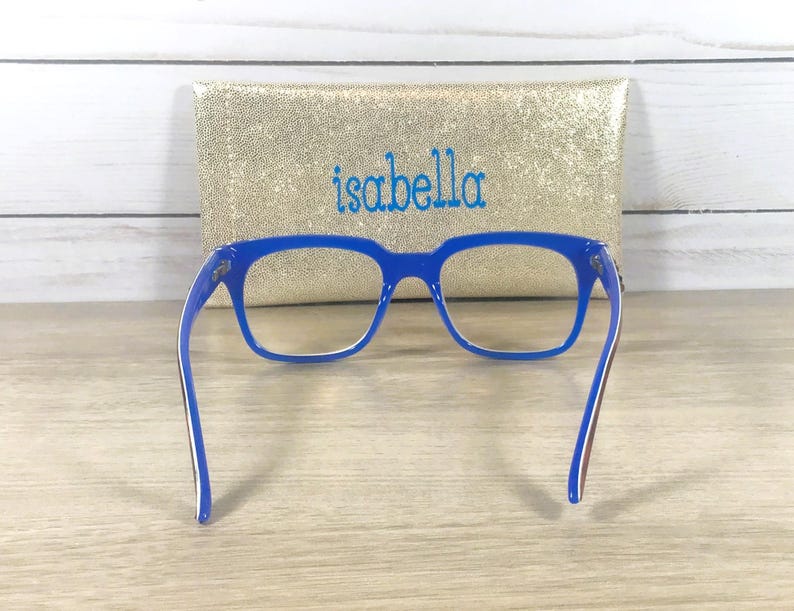 Puede incluir: Gafas azules con montura rectangular frente a un estuche dorado con lentejuelas bordado con el nombre "isabella" en azul. Las gafas tienen detalles blancos y marrones en las patillas.