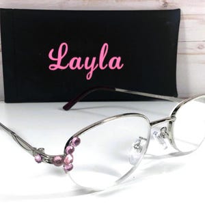May include: Silver-framed eyeglasses with clear lenses and pink gemstone accents. The glasses have a black case with the name "Layla" in pink script. The overall design is elegant and stylish.