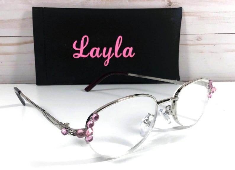 May include: Silver-framed eyeglasses with clear lenses and pink gemstone accents. The glasses have a black case with the name "Layla" in pink script. The overall design is elegant and stylish.