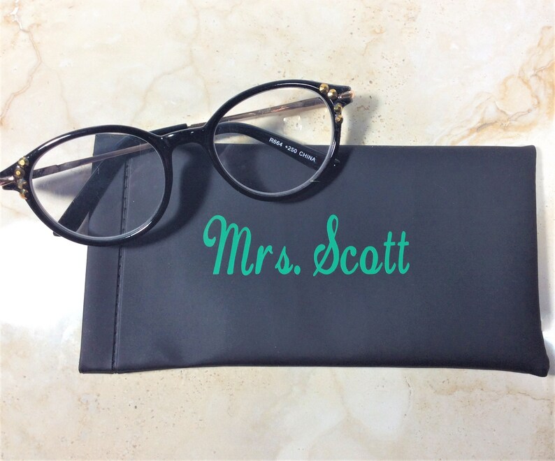 Pu&ograve; includere: Occhiali neri con montatura a occhi di gatto e dettagli dorati appoggiati su un astuccio nero. L'astuccio reca la scritta "Mrs. Scott" in verde. Gli occhiali hanno lenti trasparenti e una sottile montatura in metallo.